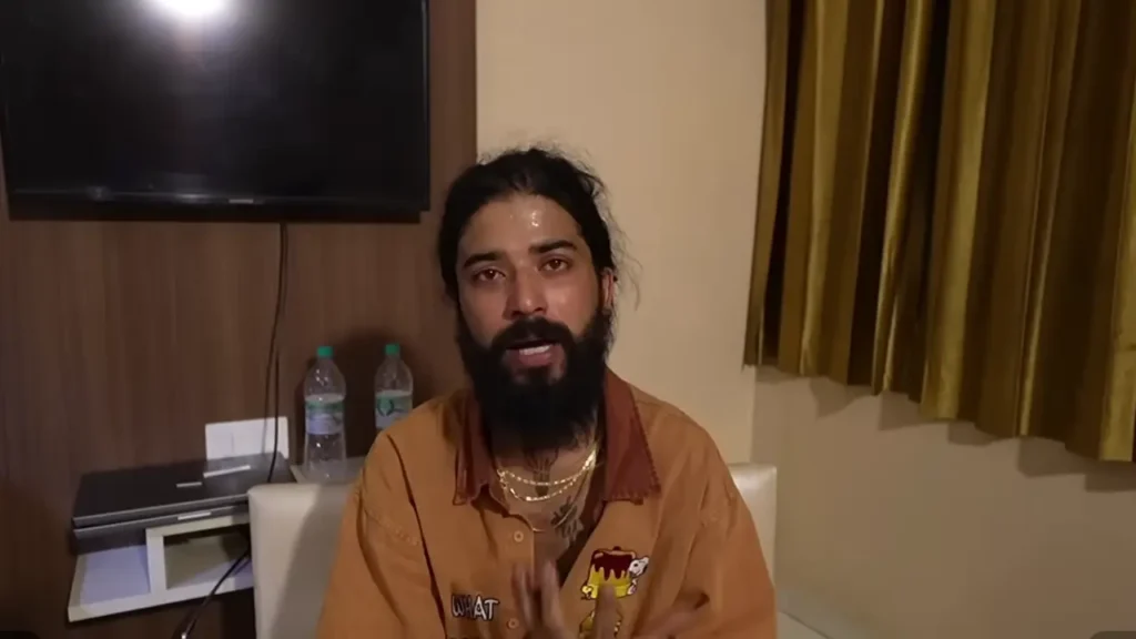 Anurag wearing an orange-brown shirt with graphic patches and gold chain necklaces, speaking in what appears to be a hotel room.