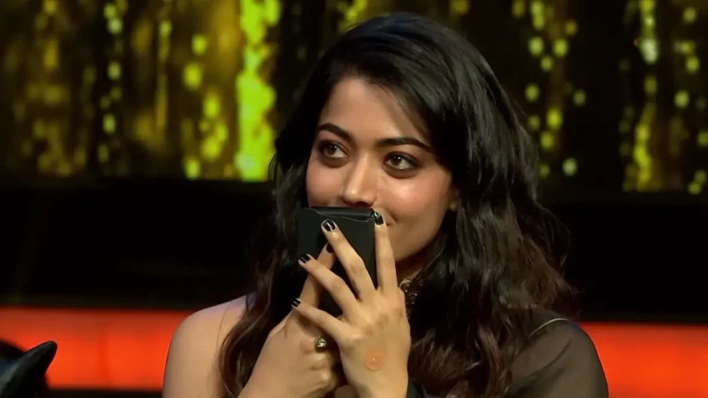 Rashmika Mandanna smiles shyly while holding a phone near her face during a talk show appearance, with warm studio lights in the background.

