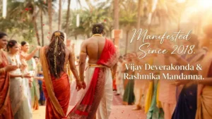A couple dressed in traditional South Indian wedding attire walk hand-in-hand down a festive aisle as guests shower flower petals, with the text “Manifested Since 2018 – Vijay Deverakonda & Rashmika Mandanna” on the right.