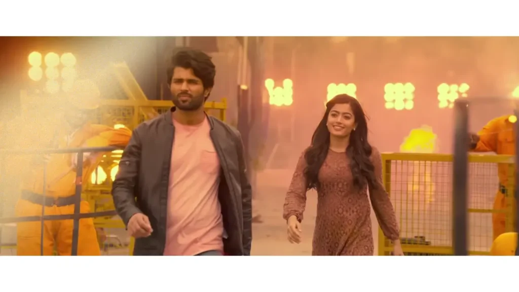 Vijay Deverakonda and Rashmika Mandanna walk confidently through a fiery industrial backdrop in a dramatic film scene.
